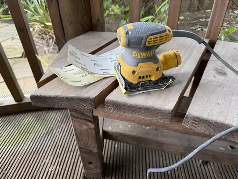 Orbital sander and strips of 80-grit abrasive paper on a timber bench seat inside a gazebo