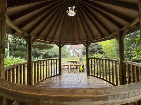 View out from inside a circular gazebo with wet decking, as a decorator jet washes a table and benches on the lawn