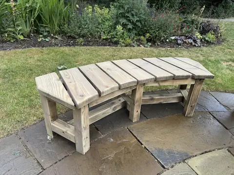 Curved slatted timber bench, sanded back to bare wood, on wet paving beside a lawn