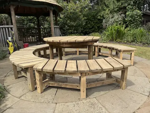 Sanded round timber table encircled by curved benches on a patio, with a pressure washer by the gazebo