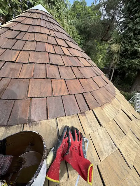 Gazebo shingle roof with the upper rows stained reddish brown and the lower rows still bare, beside a tin and gloves