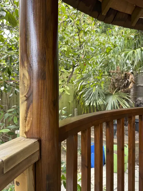 Varnished round gazebo post and curved spindle rail in rich brown next to a pale rail still to be coated
