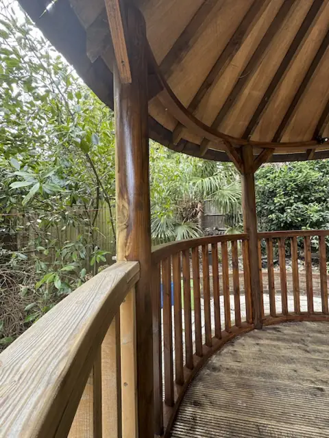 Inside a gazebo, varnished brown spindles and post beside a pale bare handrail, above grooved decking