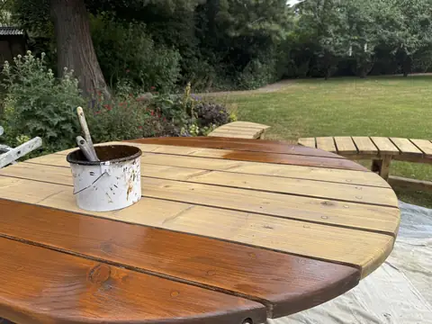 Round slatted garden table with outer slats varnished brown and middle slats bare, with a paint kettle and brushes