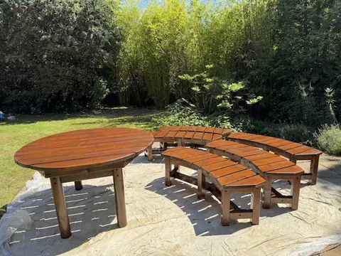 Round timber table and four curved benches varnished rich brown, on dust sheets in a sunny garden