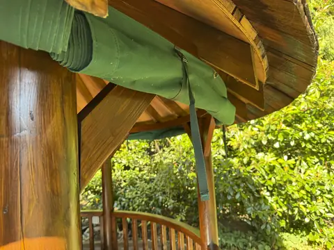 Varnished gazebo post and roof brace beneath a rolled-up green canvas curtain