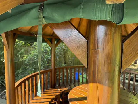 Inside a gazebo: varnished post and braces, curved spindle rail and table top under a rolled green curtain