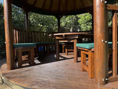 Sunlit gazebo interior with varnished posts, decking, round table and benches with green cushions